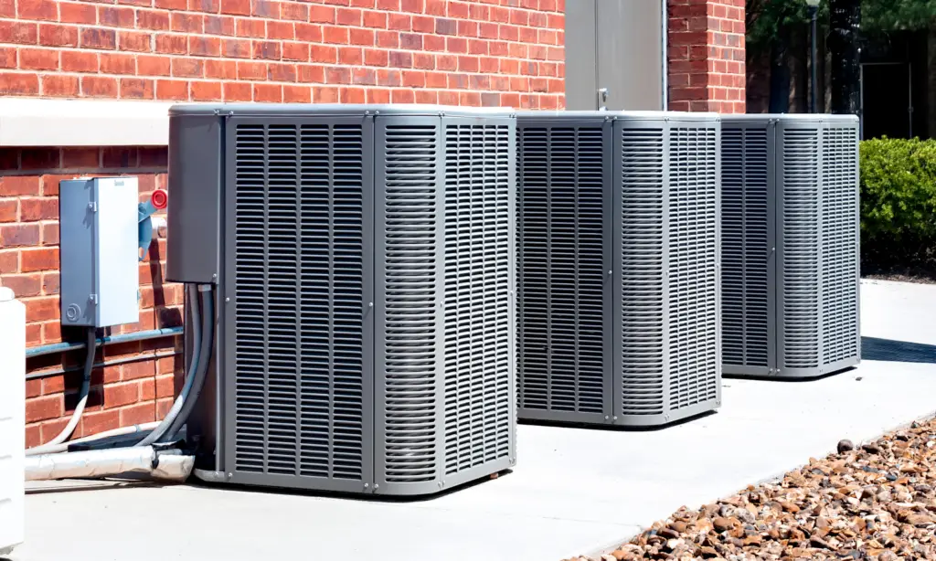 Three outdoor air conditioning units on a concrete sidewalk next to a red brick wall, with gravel on the side and greenery in the background.