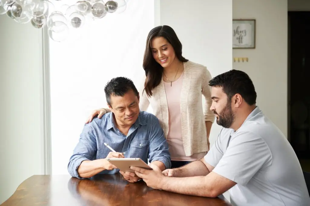 A woman and two men sitting at a table, looking at a tablet device, with the woman smiling and standing behind them.