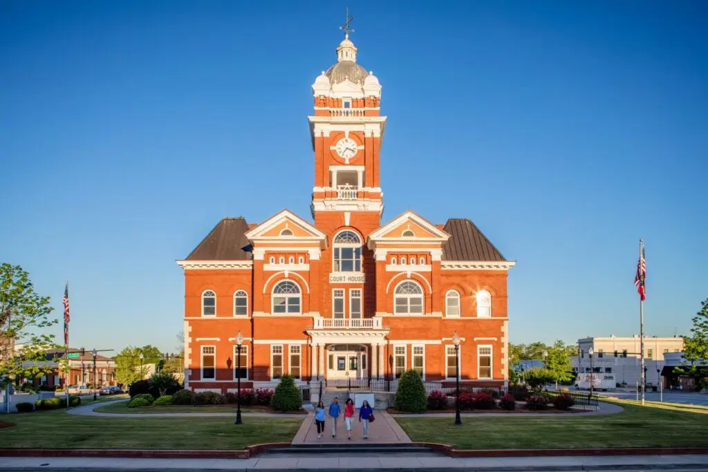 Front view of a historic red brick courthouse with a tall clock tower, surrounded by a well-maintained lawn and four women walking towards the entrance. Clear blue sky in the background.