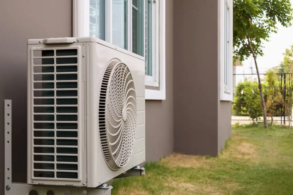An outdoor air conditioning unit installed beside a house wall with a window, on a grassy area with trees and a fence in the background.