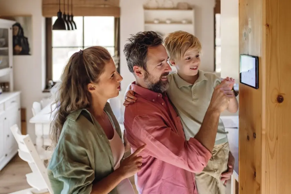A family of three, a mother, father, and young son, gathered around a tablet mounted on a wooden wall in a bright kitchen, smiling and engaging with the device.