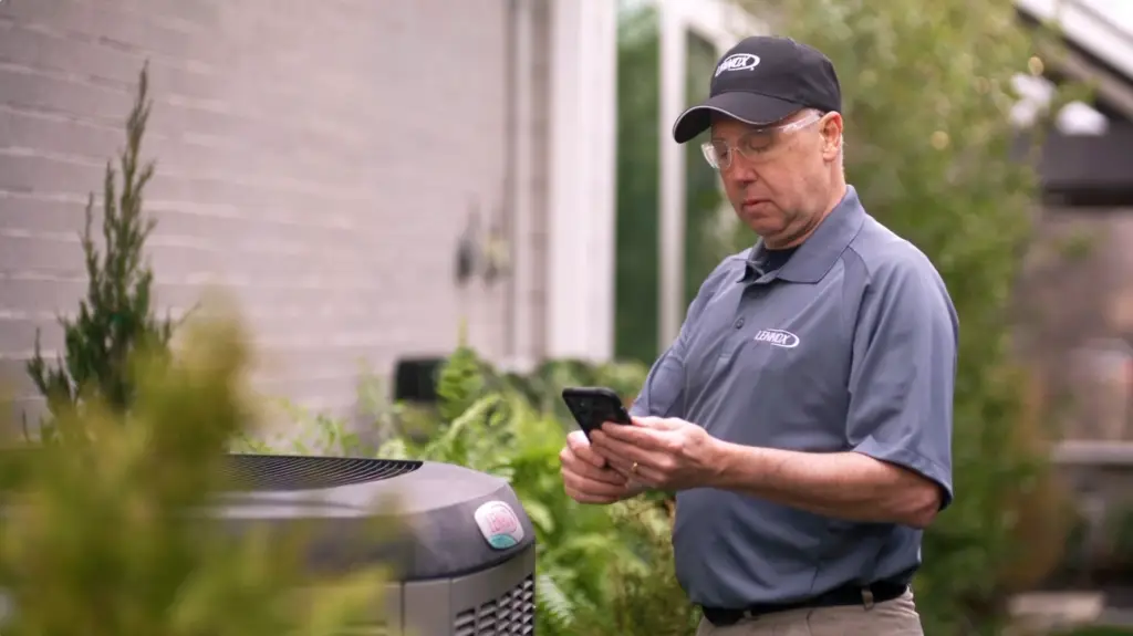 A man in a gray work shirt and black cap wearing safety glasses, holding a smartphone outdoors near an HVAC unit with greenery in the background.