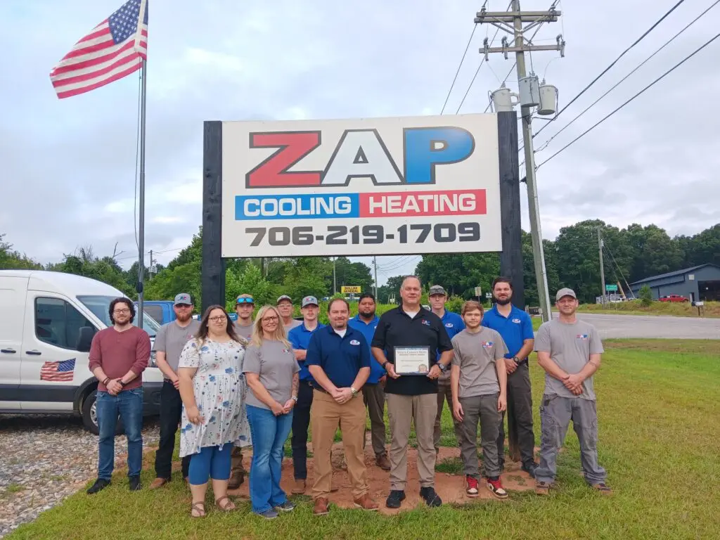 Group of people standing in front of a sign for ZAP Cooling Heating, with an American flag and utility pole nearby.