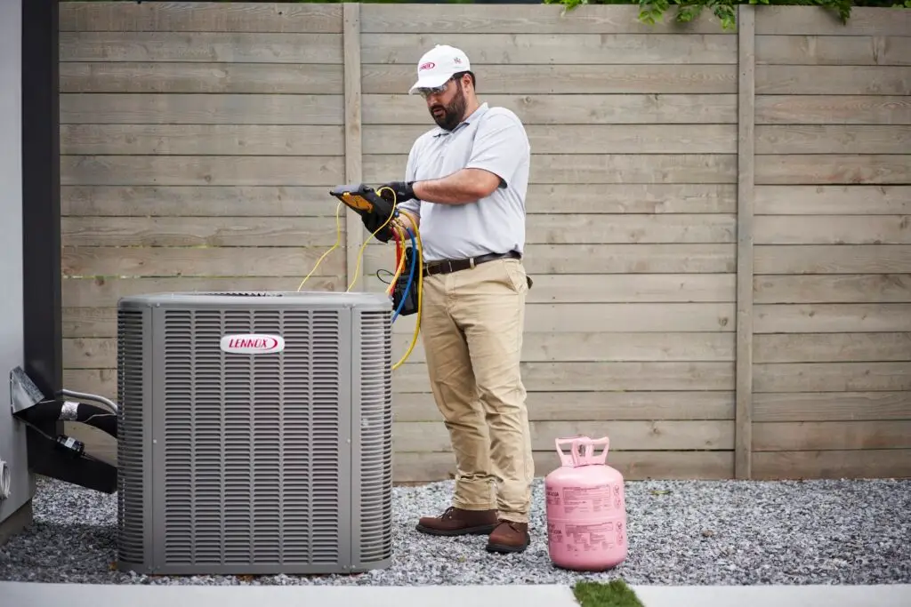 A technician wearing a white polo shirt, beige cargo pants, and protective glasses is standing outdoors, inspecting or repairing a Lennox air conditioning unit. He holds a diagnostic device connected to the unit with yellow, blue, and red wires. A pink refrigerant tank is on the gravel ground beside him, with a wooden fence and some greenery in the background.