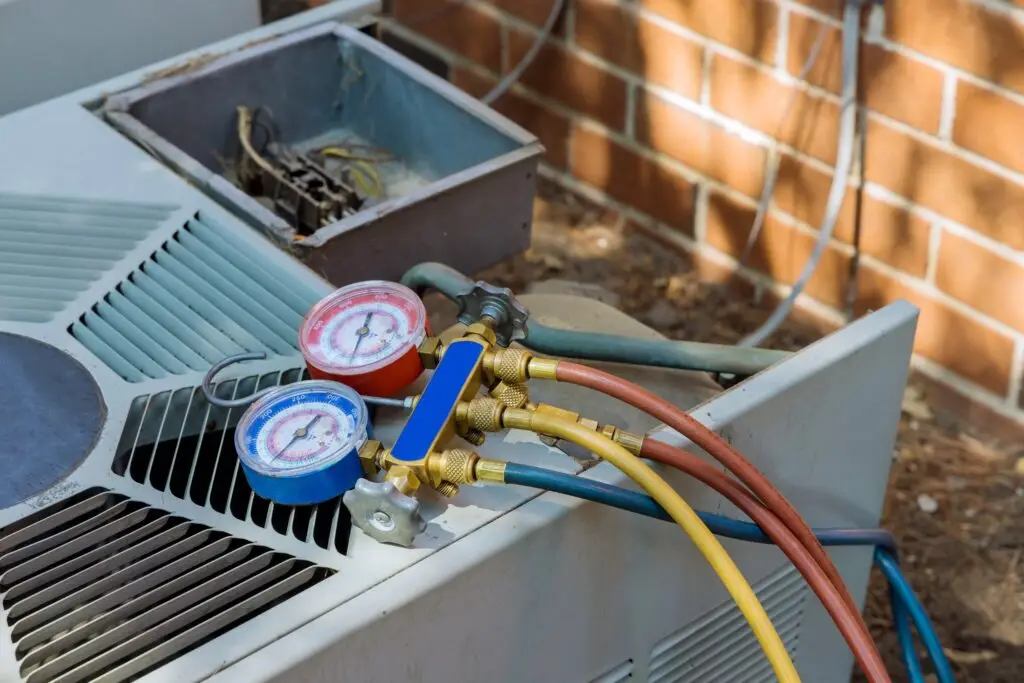 HVAC refrigeration service gauges connected to a unit with hoses, with an open electrical box nearby, against a brick wall.
