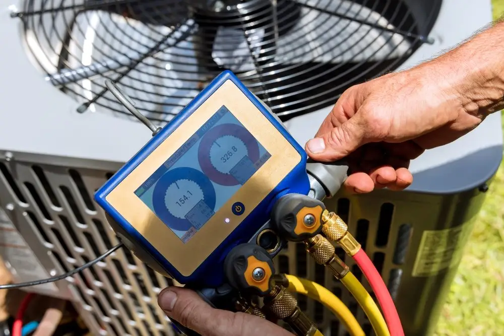 Technician holding a digital HVAC refrigerant pressure gauge and hoses connected to an air conditioning unit.