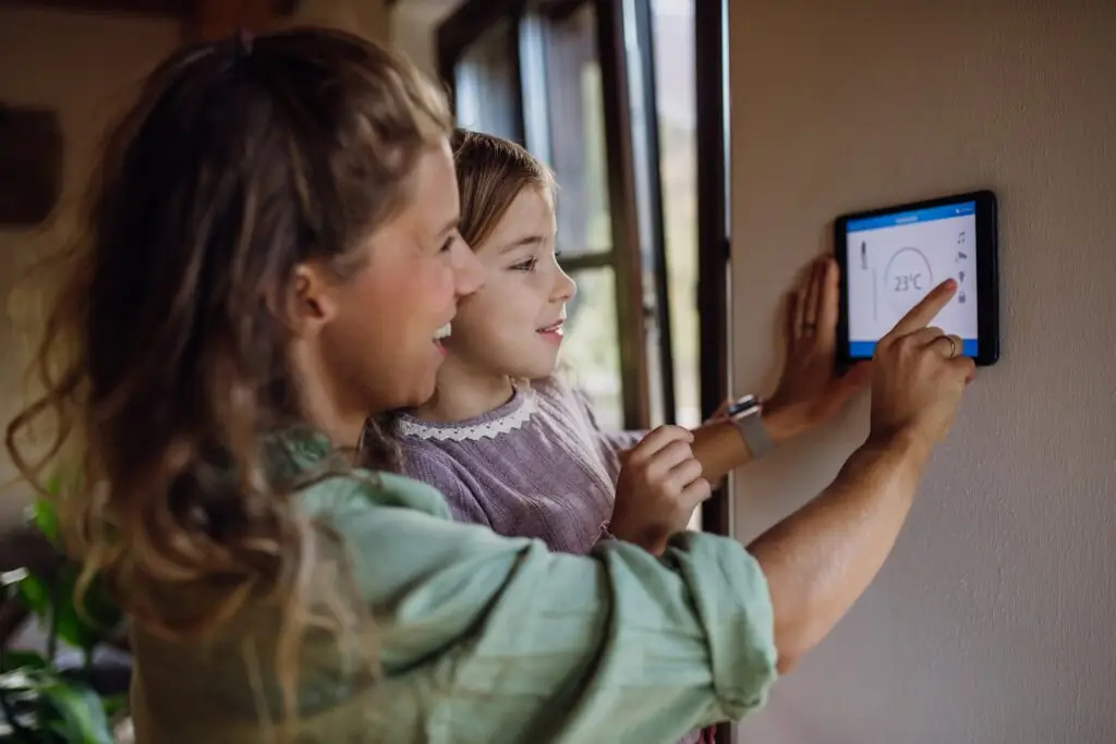 A woman and a girl adjusting a smart thermostat on the wall, the woman is pointing at the screen showing 23°C.